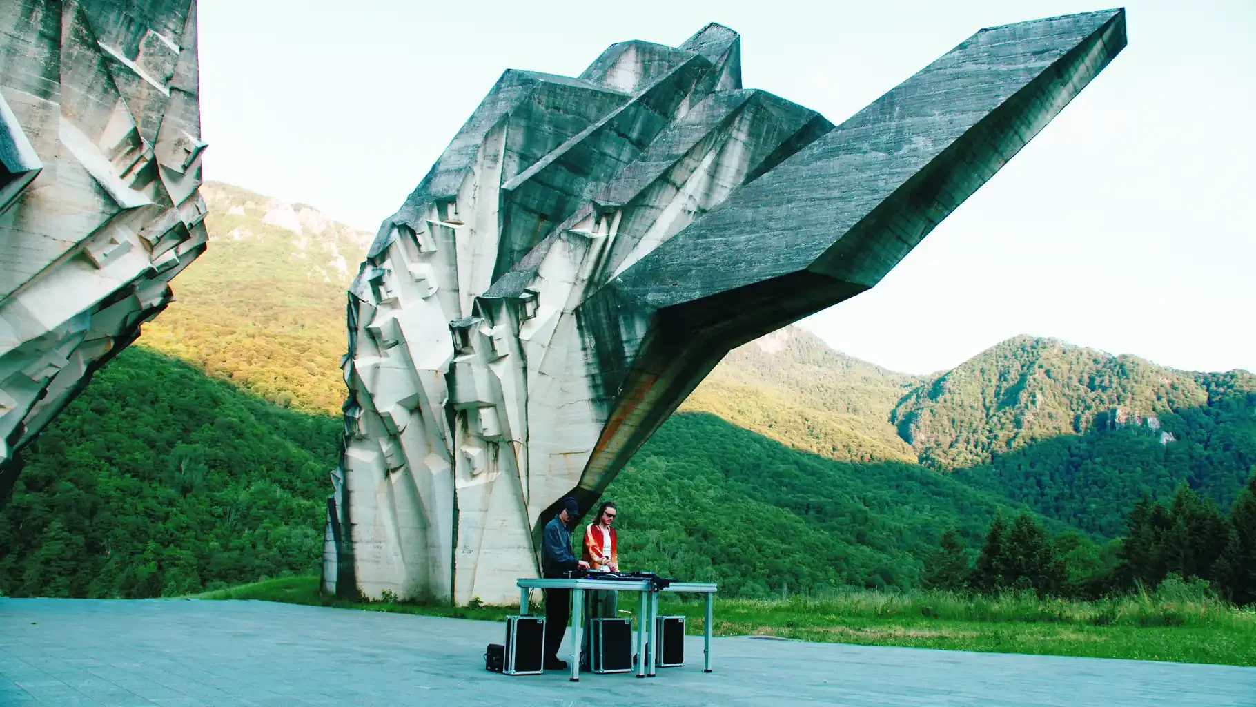 DJ duo performing beneath monumental sculptural concrete architecture in green mountains
