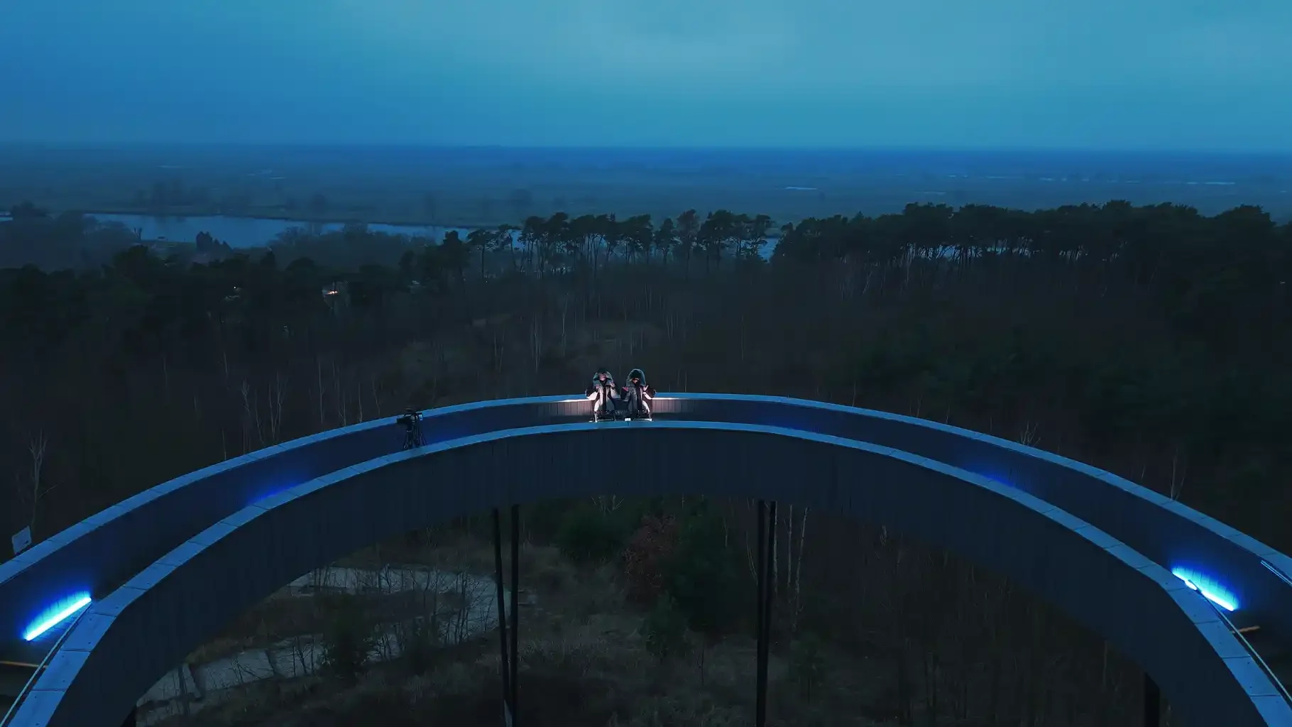 Aerial view of two DJs performing on a curved elevated walkway above a forest